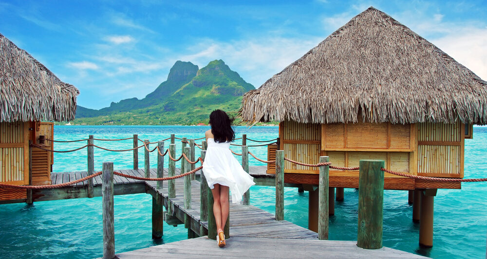 woman in a white flowy dress leaning against the side of an overwater bungalow in Bora Bora with green mountain and blue water in front of her