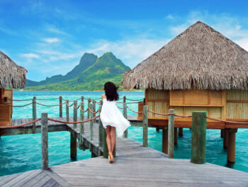 woman in a white flowy dress leaning against the side of an overwater bungalow in Bora Bora with green mountain and blue water in front of her