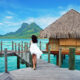 woman in a white flowy dress leaning against the side of an overwater bungalow in Bora Bora with green mountain and blue water in front of her