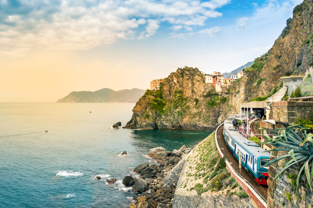 A train passing along the coastline in Cinque Terre Italy with train track carved out of the cliff with ocean views 