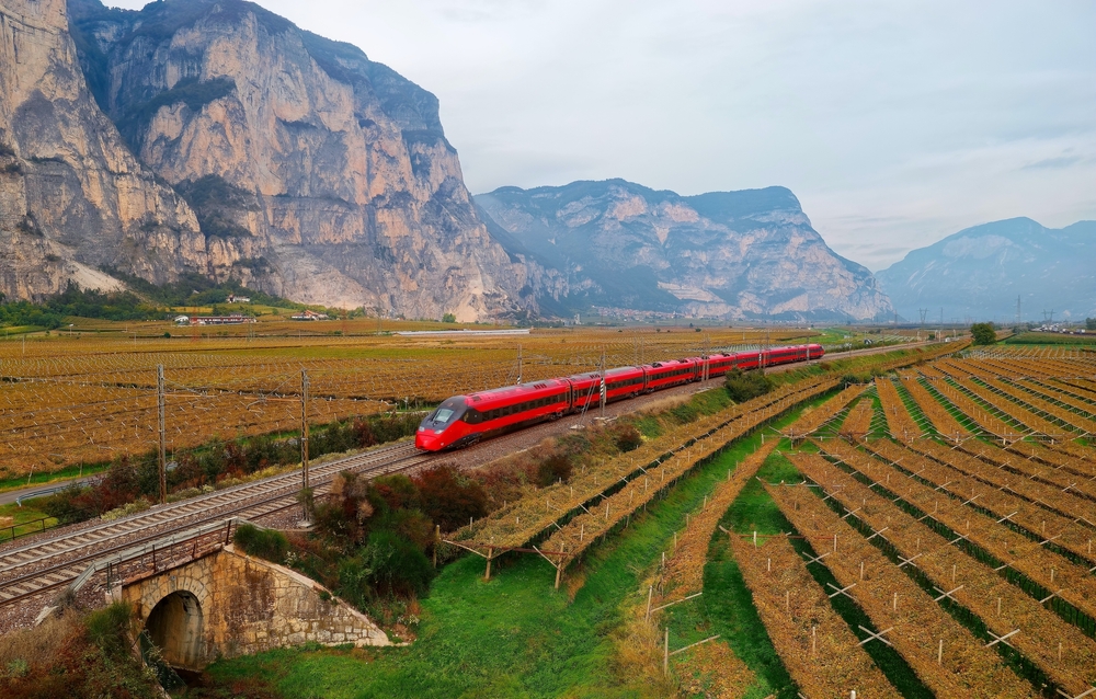 One of the red fast trains going through Italian farm fields with mountains in the background on way to Florence