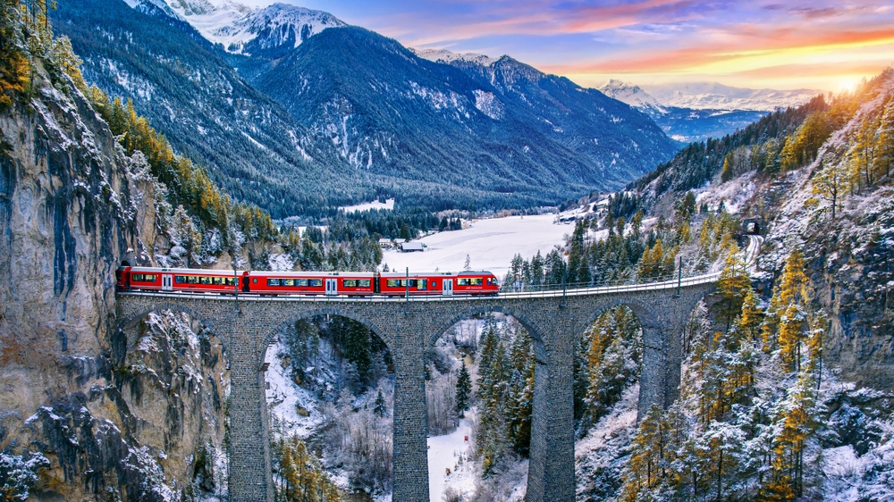 a red train passing over a viaduct in Switzerland at sunset with snow-capped mountains and a lake