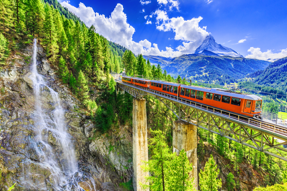 a train passing over a bridge with a waterfall and mountain in the background surrounded by lush green trees on one of the 