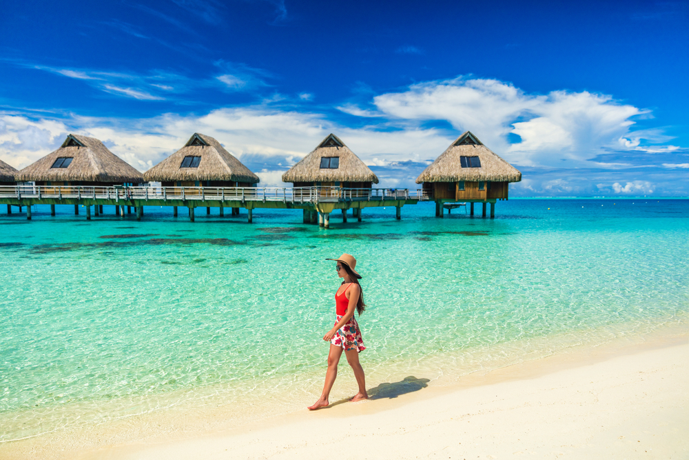 woman walking on the beach in a spaghetti strap top, sun hat, and short skir, theres over the water bungalows in the background