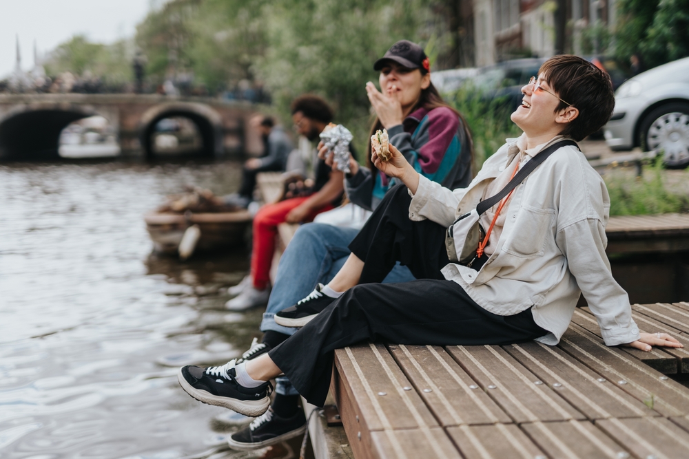 A group of friends sit on the edge of the water knowing what to wear to Amsterdam in spring, as they eat and are comfortable in their pants, sneakers, and button down shirts. 