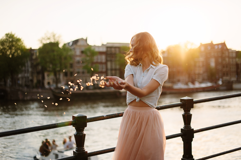 A woman poses on the bridge by the water, her hands cupped tougher as petals drift from them: she wears a midi skirt and a white button down. 