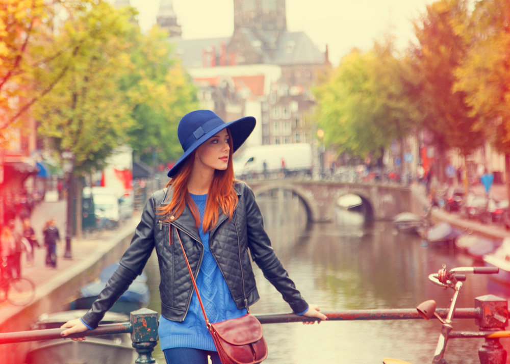 A woman poses on the water, in full street style, know what to wear to Amsterdam which includes jeans, a sweater, a leather jacket and a blue hat. 