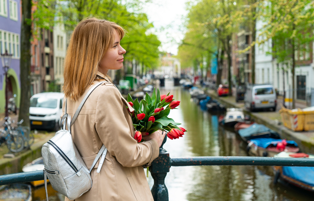 A woman with a small silver backpack and beige trench coat clutches tulips and looks off camera in Amsterdam. 