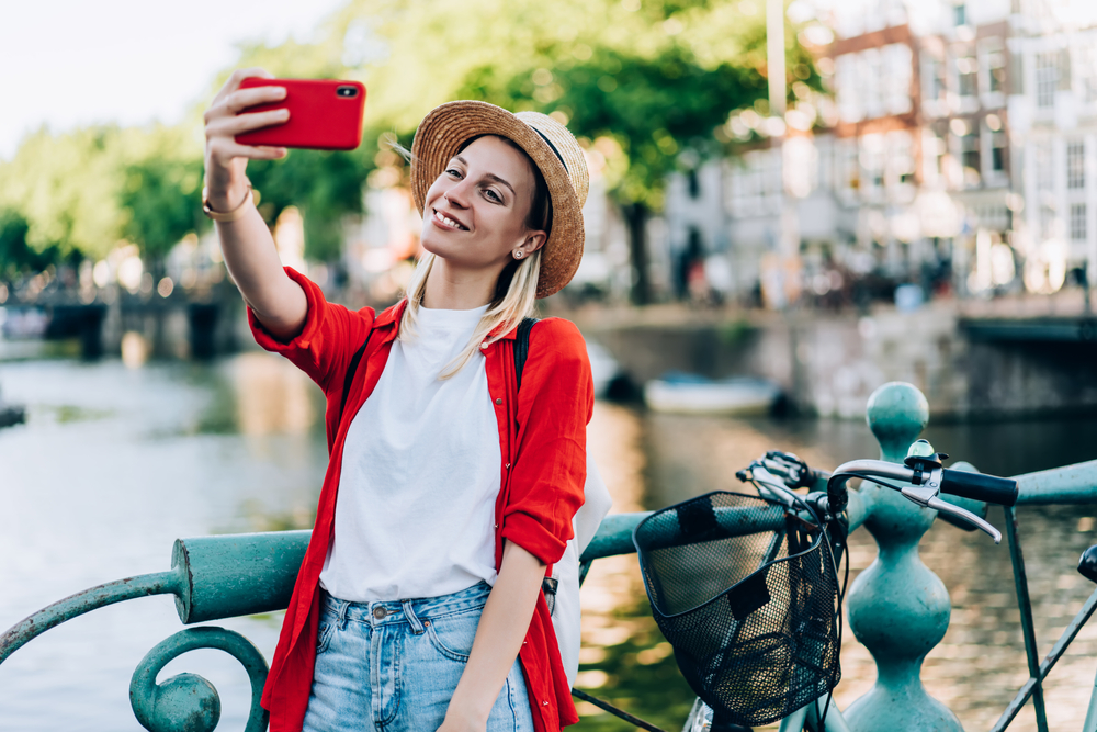 A woman in a white t-shirt and red long sleeve shirt smiles for a selfie on the water: her red phone case matches her shirt and contrasts her jeans. 