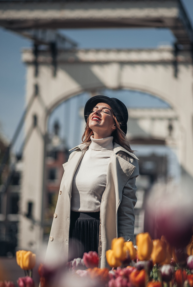 A woman knows what to wear to Amsterdam in the spring, as she enjoys the warmth as she turns her face to the sun while wearing a light turtle neck, skirt and a trench coat. 