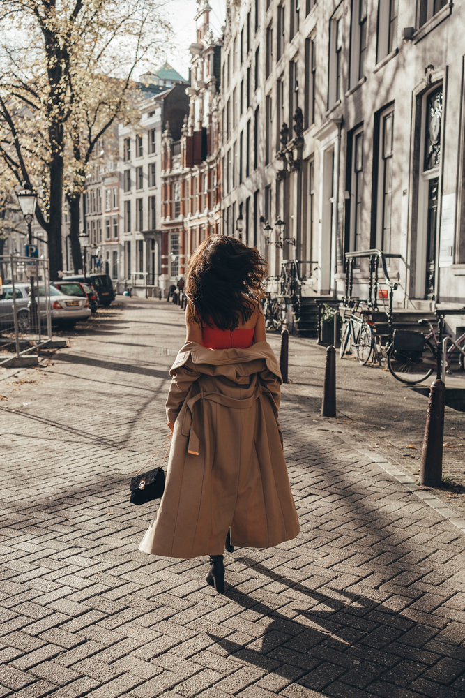 A woman knows what to wear to Amsterdam in spring as she walks the pathways of the street in a orange tank and a trench jacket sliding off her shoulders. 