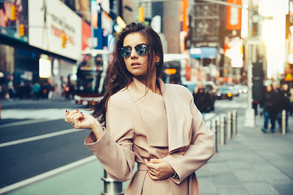 A model poses in Times Square with a long, classic trench coat on to keep her warm. She wears a pair of oversized sunglasses as well. 