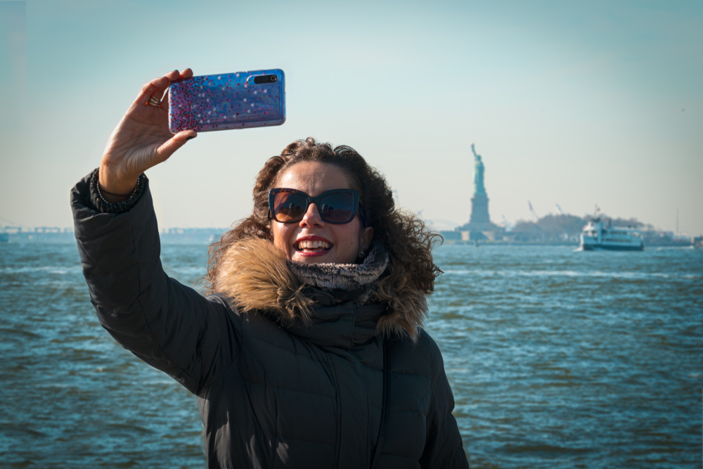 When wondering what to wear in New York City in winter, channel this girl as she wears a faux fur parka, sunglasses and a neck gaiter as she takes a selfie with the statue of Liberty in the background. 