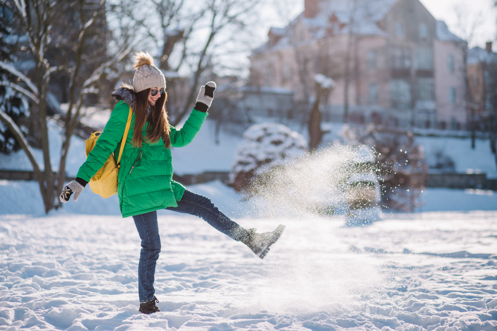 A woman enjoys snow in Central Park as she is wearing her beanie, boots, jeans, gloves and a green puffer jacket. 