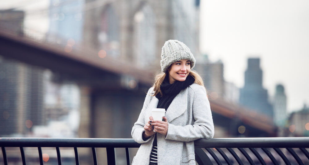 a woman smiling in front of a bridge in NYC wearing a grey beanie hat, a black scarf, and a white jacket leaning against a railing.