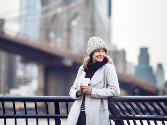 a woman smiling in front of a bridge in NYC wearing a grey beanie hat, a black scarf, and a white jacket leaning against a railing.