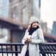 a woman smiling in front of a bridge in NYC wearing a grey beanie hat, a black scarf, and a white jacket leaning against a railing.