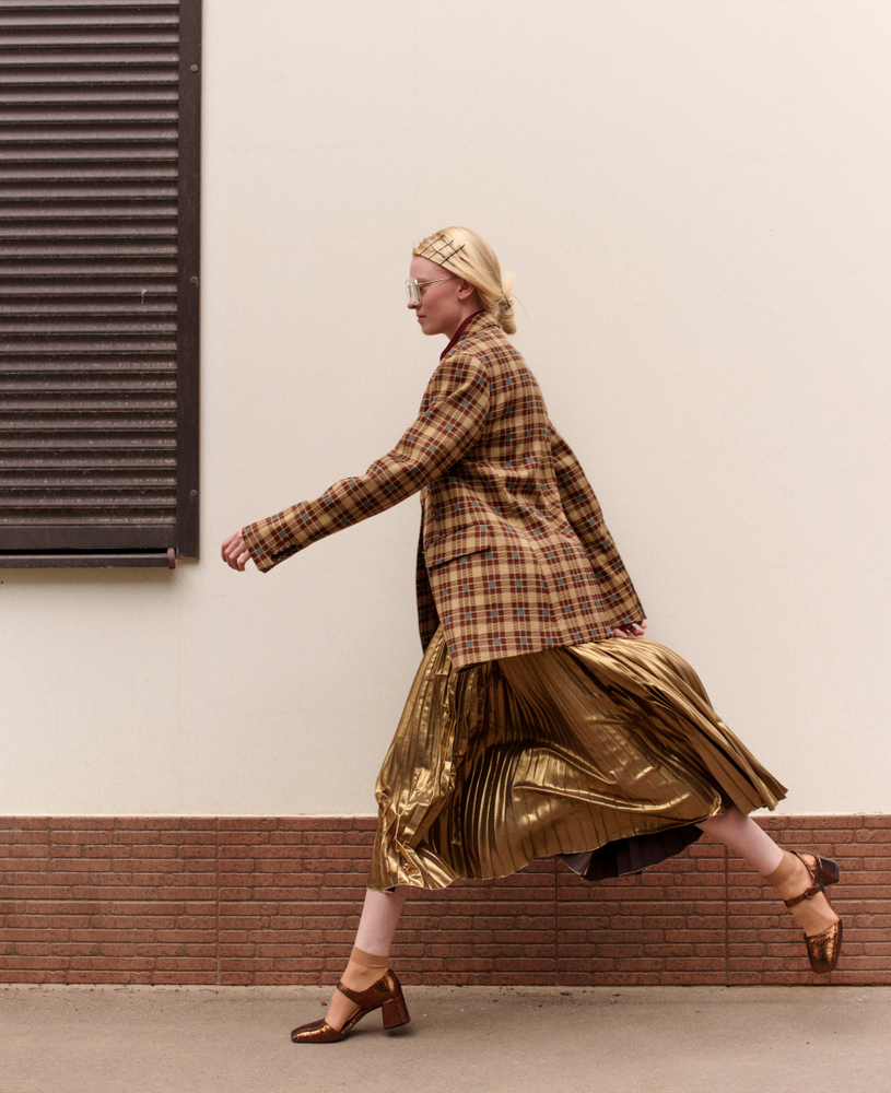 A woman walks down the street in New York with a gold midi skirt on, gold heels, and a classic plaid jacket. 