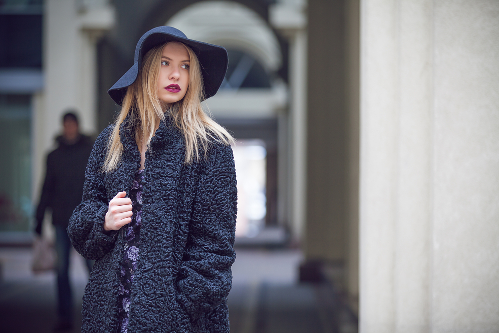 A woman explores NYC in a large floppy hat, a fuzzy coat, and a knit sweater, all in hues of blues and purples, which match her purple lipstick as well. 