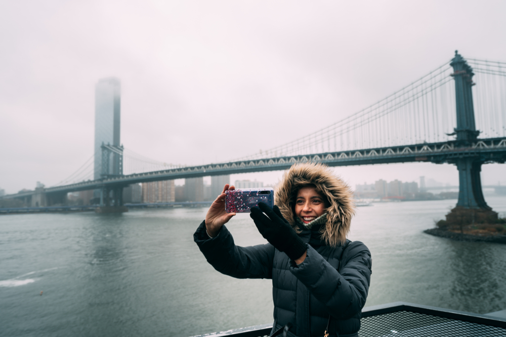 A woman knows what to wear in New York City in winter, as she takes a selfie with the Brooklyn bridge: her faux fur parka and gloves keep her toasty. 