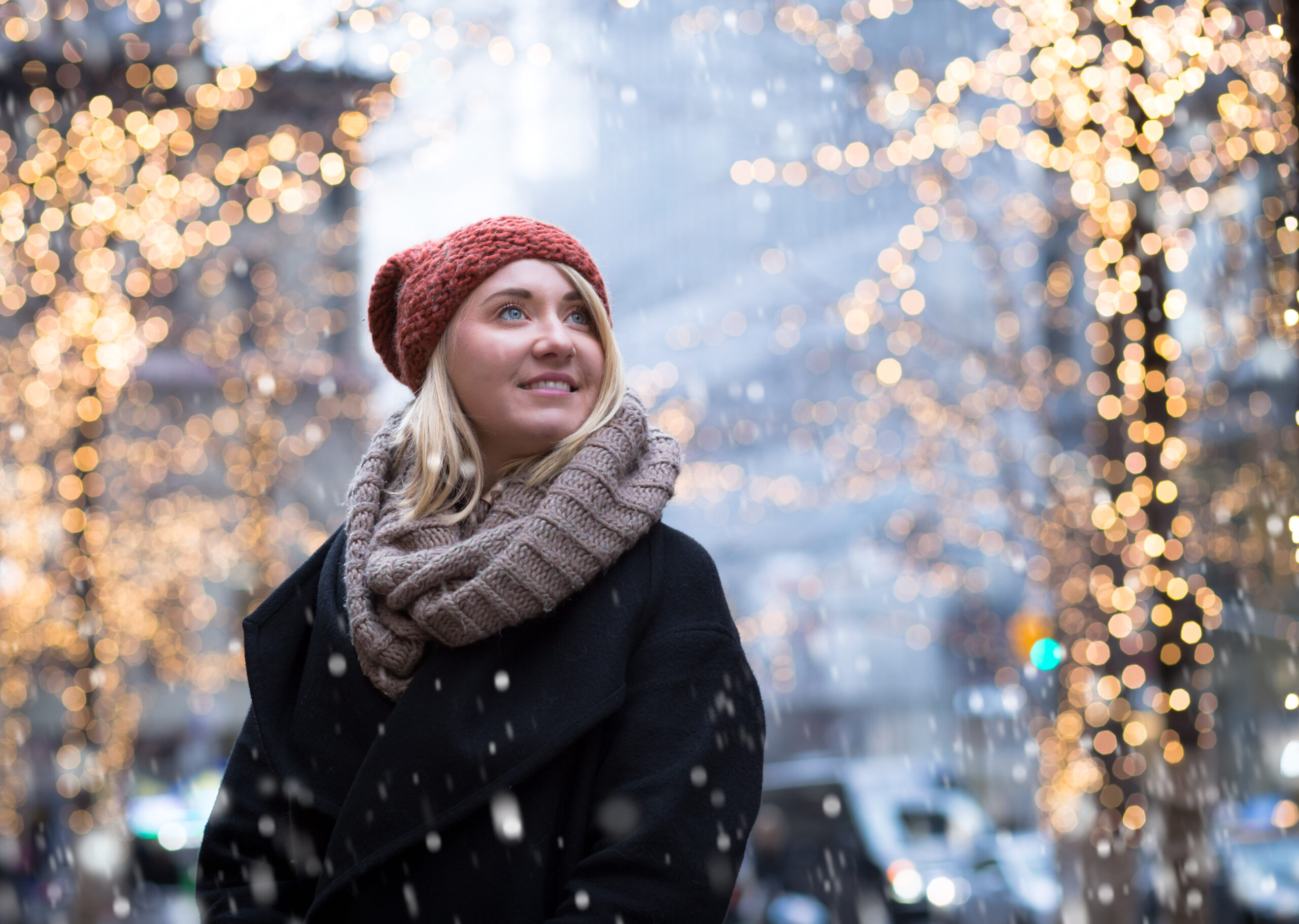 A woman in a thick knit scarf and coat and beanie watches snow fall around her while Christmas lights are blurred in the background of the city. 