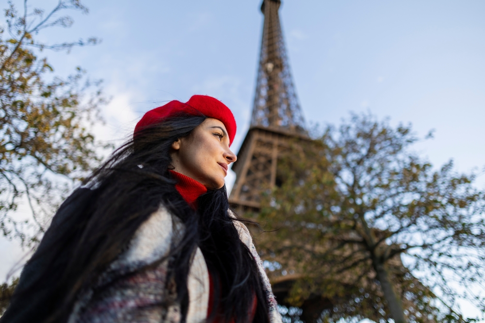 A woman in a red beret, turtleneck and wool jacket stands below the Eiffel Tower, knowing exactly what to wear in Paris in winter. 