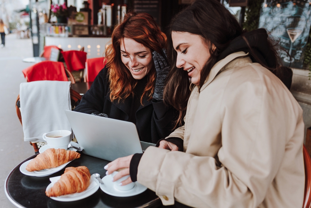 Two friends sit at a cafe with coffee and croissants, enjoying the cool weather in Paris in winter  with their gloves and trench coats on. 