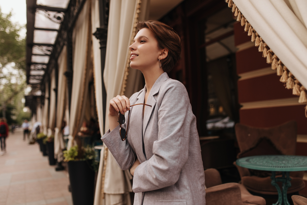 A woman knows what to wear in Paris in winter as she leans against a cafe edge wearing a thicker grey blazer: she holds sunglasses in her right hand. 