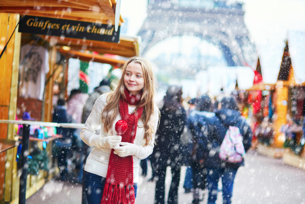 A young blonde girl examines a Christmas market by the Eiffel Tower as it snows: she wears gloves, a sweater and a scarf. 