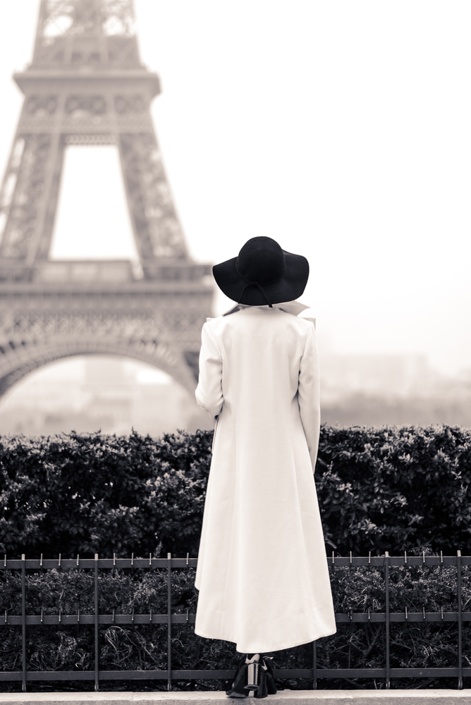 In a black and a white shot, a woman faces the Eiffel Tower while wearing a black hat and a long, white trench coat. 