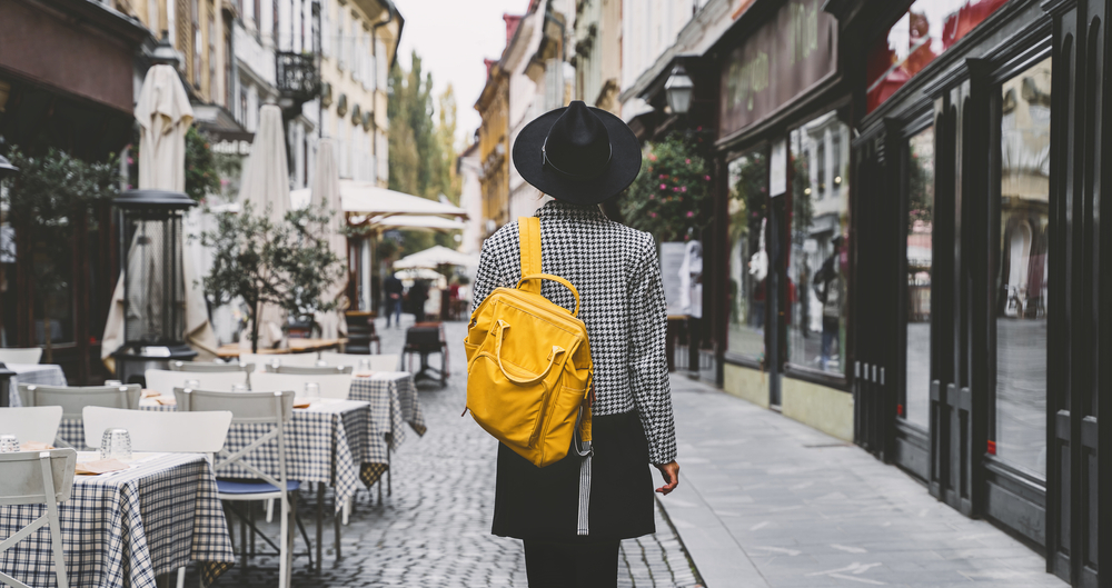 A woman in a skirt, tights, and tweed-like jacket walks down cobblestone roads in France while wearing a yellow backpack and a black hat. 