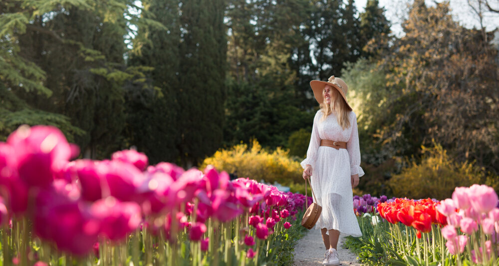 A woman walks down a pathway, knowing what to wear to Amsterdam in spring, as she wears a white long sleeve midi dress with a belt, hat, and matching purse, perfect for the warmer months like May!