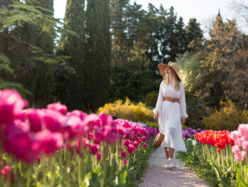 A woman walks down a pathway, knowing what to wear to Amsterdam in spring, as she wears a white long sleeve midi dress with a belt, hat, and matching purse, perfect for the warmer months like May!