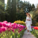 A woman walks down a pathway, knowing what to wear to Amsterdam in spring, as she wears a white long sleeve midi dress with a belt, hat, and matching purse, perfect for the warmer months like May!