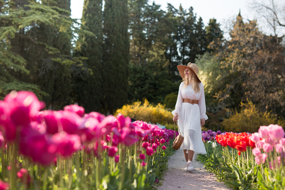 A woman walks down a pathway of pink tulips in a white midi dress that is long sleeves. She wears a belt, a sun hat, and a woven bag that all matches! 