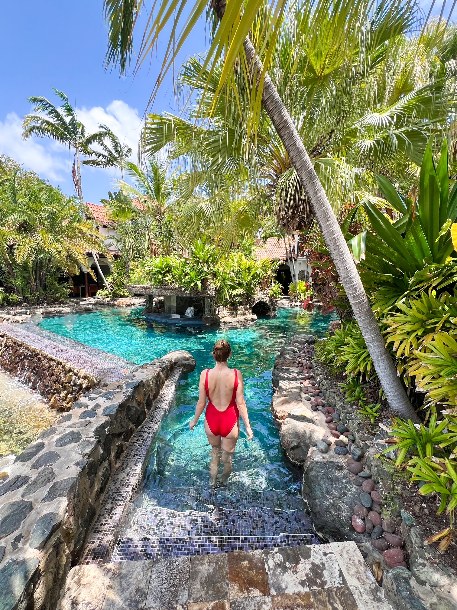 woman in red bathing suit with her hair tied up walking into a beautiful blue pool that is surrounded by trees and a beautiful rock oasis 