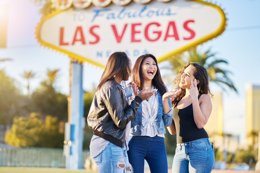 A group of three friends laugh with one another in front of the Vegas sign, all wearing jeans and tank tops and jackets, ready and knowing what to wear in Las Vegas. 